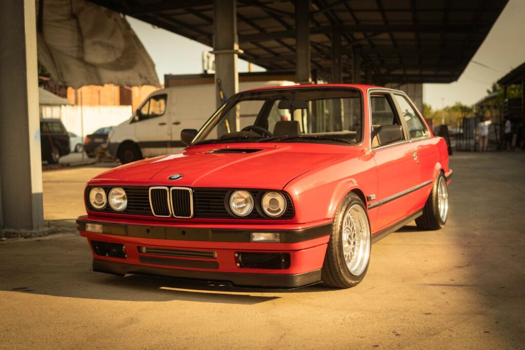 A classic red BMW E30 coupe parked in an industrial setting under warm sunlight.